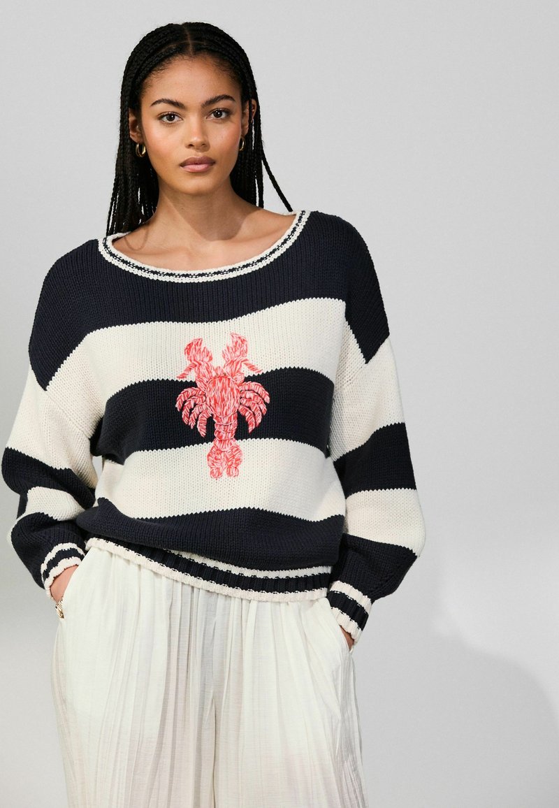 Femme aux cheveux tressés portant un pull à rayures noir et blanc avec un motif de homard rouge et un pantalon blanc à jambes larges, mains dans les poches.