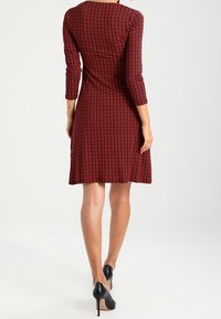 Robe rouge avec un motif géométrique, manches trois-quarts et taille cintrée. Tissu lisse, longueur genou, portée avec des talons noirs.