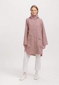 Ljusrosa vattentät regnjacka med huva, två framfickor och lös passform, kombinerad med vita byxor och sneakers.