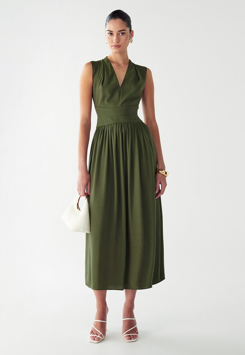 Vestido sin mangas verde oliva con escote en V, cintura ajustada y falda fluida, combinado con un bolso blanco y sandalias blancas de tiras.
