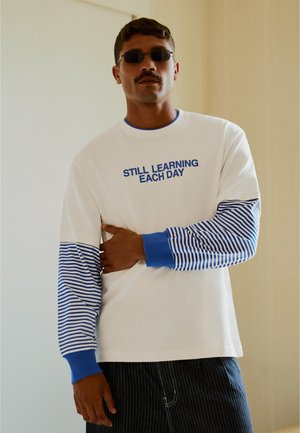 Muž s knírkem a slunečními brýlemi, který nosí bílou košili s dlouhými rukávy s modrými pruhy a nápisem "STÁLE SE KAŽDÝ DEN UČÍM."