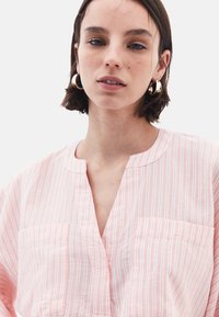 Camicia a righe rosa con scollatura a V e maniche corte, dotata di due tasche sul petto e in un tessuto leggero e testurizzato. Orecchini circolari visibili.