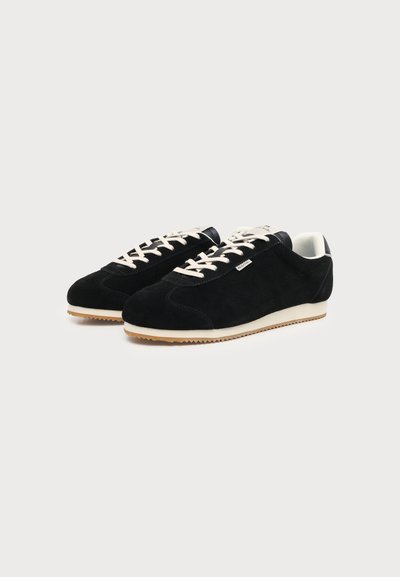 Ζευγάρι μαύρα σούεντ sneakers χαμηλού ύψους με λευκά κορδόνια, λευκή εσωτερική επένδυση και μπεζ λαστιχένιες σόλες σε ανοιχτό γκρι φόντο.