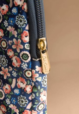 Złoty suwak z wygrawerowanym napisem "Cath Kidston" na niebieskiej tkaninie w kwiaty z różowymi, białymi i zielonymi kwiatami na beżowym tle.