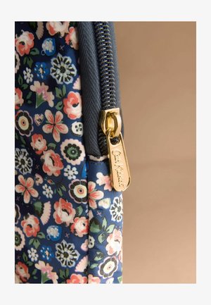 Złoty suwak z wygrawerowanym napisem "Cath Kidston" na niebieskiej tkaninie w kwiaty z różowymi, białymi i zielonymi kwiatami na beżowym tle.