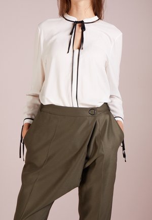 Frau trägt eine weiße Bluse mit schwarzem Besatz und Bindebändern, kombiniert mit asymmetrischen, olivgrünen, weit geschnittenen Hosen mit einer vorderen Schnalle.