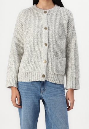 Žena v světle šedém pleteném kardiganu s předními kapsami a stříbrnými knoflíky, v kombinaci s modrými džínami na bílém pozadí.