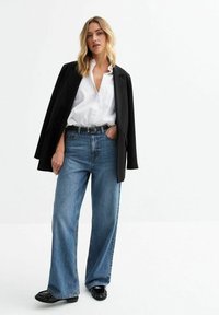 Szerokie niebieskie dżinsy połączone z białą koszulą zapinaną na guziki i czarnym żakietem, podkreślone czarnym paskiem i błyszczącymi czarnymi butami.