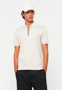 Beige piké skjorte med korte ermer, glidelås i kragen og svak stripe detalj ved glidelåsen. Kombinert med mørke shorts og en caps.