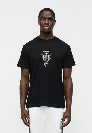 Man wearing black Versace Jeans Couture t-shirt and white pants with black designs, standing against plain white background.