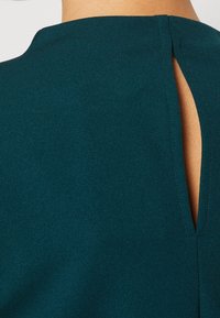 Tyrkysový top z jemné tkaniny s vysokým výstřihem a vetracím detailem na zadní straně, který ukazuje hladkou strukturu a čisté linie.