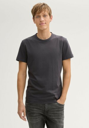 Camiseta de cuello redondo en gris oscuro, hecha de algodón. Mangas cortas y un diseño ajustado. Modelo de pie con las manos en los bolsillos, vistiendo jeans oscuros.