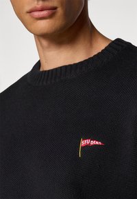 Svart strikket genser med ribbet rund hals og et lite brodert rødt og gult "STUDENTS" flaggdetalj på brystet.