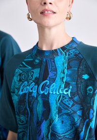 Kurze Ärmel, türkis- und schwarz gemusterte Bluse mit Rundhalsausschnitt und gesticktem "Carlos Colucci"-Logo in Blau. Strukturierte Stoffdetails.