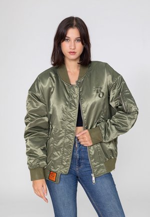 Jeune femme portant une veste bomber vert olive brillante par-dessus un crop top noir et un jean bleu, debout devant un fond gris uni.