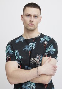 Černé tričko s krátkým rukávem s barevným tropickým vzorem palmového listu; model má na zápěstí stříbrný náramek a kulatý výstřih.