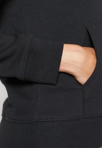 Felpa nera con tasca sul davanti. Presenta polsini e orlo a costine, con dettagli di cucitura visibili. Texture morbida.