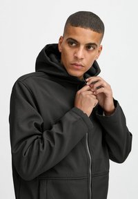 Veste à capuche noire avec fermeture éclair à l'avant, présentant une texture lisse et un motif subtil. Comprend une coupe décontractée et des poignets élastiques.