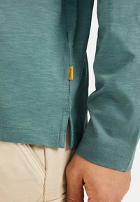 Langærmet skjorte i dæmpet teal, lavet af struktureret stof. Har en sideslids og et lille gult logo-tag nær kanten. Materiale af bomuldsblanding.