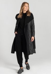 Czarny długi płaszcz o warstwowej konstrukcji, z pluszowym kołnierzem, puszystą fakturą i dużymi guzikami. W połączeniu z czarnym swetrem i spodniami.