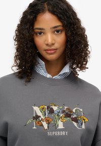 Sudadera gris con bordado floral que incluye el texto "NYC" y "SUPERDRY", complementada con una camisa de collar a rayas azul y blanca debajo.