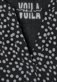 Blouse noire avec un décolleté en V, présentant un motif à pois blancs répété. Le tissu a une surface légèrement texturée et une finition lisse.