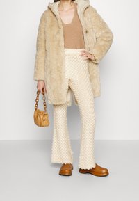 Beige fuskpälsjacka, tan stickat topp, krämfärgade strukturerade utsvängda byxor, bruna chunky skor och en liten karamellfärgad väska med kedjehandtag.