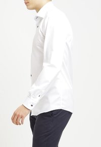 White long-sleeve dress shirt with a smooth texture, featuring black buttons on the cuffs and under the collar, tailored fit, and rounded hem.