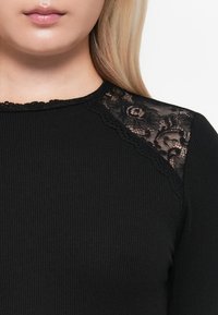 Black ribbed long-sleeve top featuring sheer lace accents on the shoulders, with scalloped detailing along the neckline.