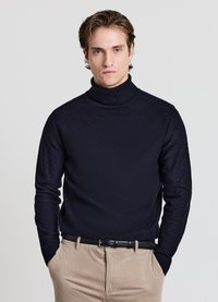 Marineblauer Rollkragenpullover mit strukturiertem Diamantmuster, gerippten Bündchen und Kragen, kombiniert mit beigen Hosen und einem schwarzen Gürtel.