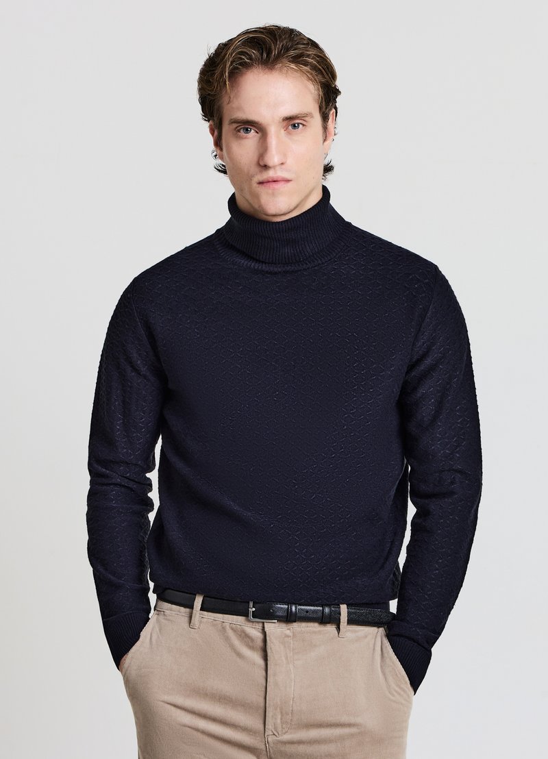 Marineblauer Rollkragenpullover mit strukturiertem Diamantmuster, gerippten Bündchen und Kragen, kombiniert mit beigen Hosen und einem schwarzen Gürtel.