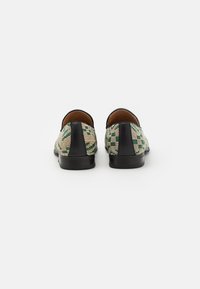 Chaussures à enfiler tissées avec un motif vert et beige, accents noirs au talon et intérieur en cuir lisse. Semelle plate noire de forme arrondie.