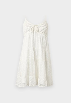 Witte mouwloze zomerjurk met gehaakte lijfje, verstelbare strik aan de voorkant en een gelaagde, getextureerde rok met kant- en stippenmotieven.