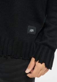 Pull en maille noir avec ourlet et poignets côtelés. Présente des bords effilochés et une petite étiquette cousue avec "Sixth June" en blanc sur fond noir.