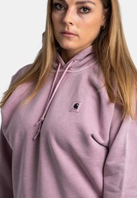 Hoodie rose clair en tissu doux et texturé, avec une capuche à cordon et un petit logo noir sur le devant.