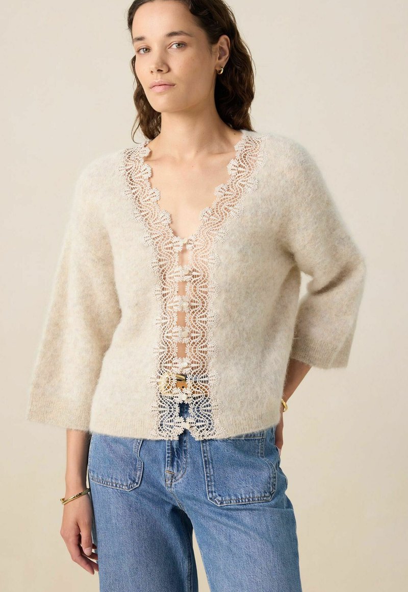 Pull en mohair beige avec un décolleté en V profond, orné de délicates bordures en dentelle blanche. Coupe ample avec des manches 3/4, associé à un jean en denim bleu.