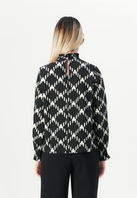 Donna con lunghi capelli ombre che indossa una blusa a maniche lunghe bianca e nera a fantasia con apertura a chiave sulla schiena e pantaloni neri su uno sfondo bianco.