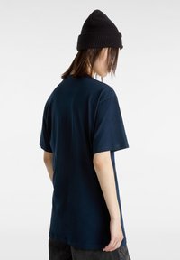 Personne aux cheveux mi-longs portant un bonnet en tricot noir et un t-shirt bleu foncé oversize, vue de dos sur un fond uni.