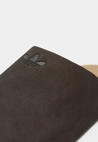 Temno rjave semišove natikače z gladko teksturo in povišanim črnim Adidasovim logotipom na zgornjem delu. Konturiran peščeni vložek vidn na strani.