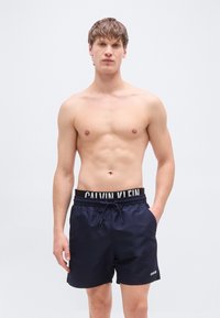 Μπλε σκούρο μαγιό σορτς με μαύρη μέση που φέρει το λογότυπο «CALVIN KLEIN». Το υλικό φαίνεται λείο με ελαφρύ σχεδιασμό και πλαϊνές τσέπες.