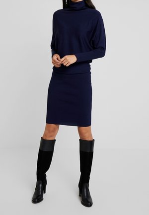 Femme portant une robe col roulé bleu marine à manches longues et des bottes noires montantes jusqu'aux genoux, debout devant un fond gris clair uni.