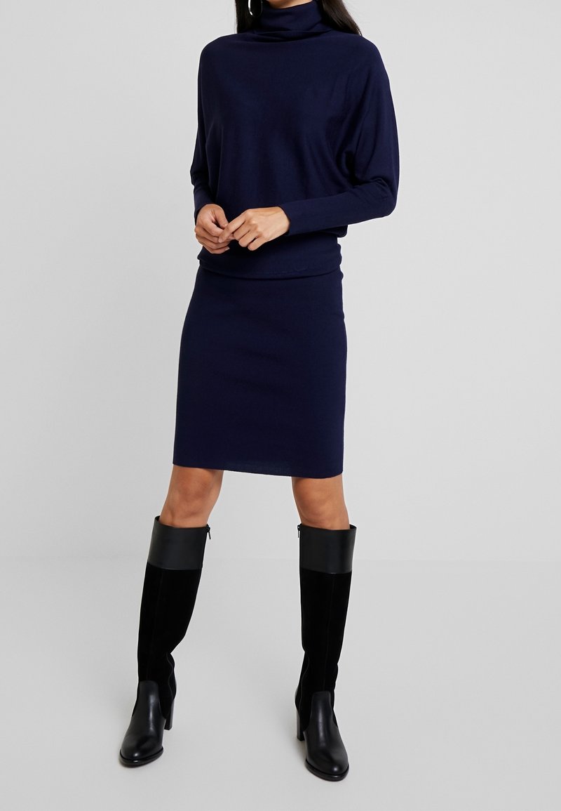 Femme portant une robe col roulé bleu marine à manches longues et des bottes noires montantes jusqu'aux genoux, debout devant un fond gris clair uni.