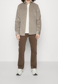 Vaaleanruskea corduroy-paita, napitettu, yhdistetty ruskeisiin housuihin. Malli pukeutunut valkoiseen t-paitaan ja harmaisiin lenkkareihin, joissa mustia yksityiskohtia.