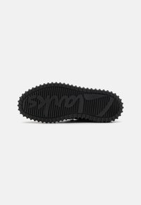 Zwarte rubberen sneakerzool met uitgesproken ribbels en een circulair patroon. De merknaam is in een vet, gebogen lettertype in het midden geperst.