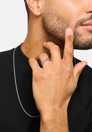 Mann mit getrimmtem Bart trägt silberne Kettenhalskette, berührt sein Gesicht mit der linken Hand und zeigt dabei einen kunstvoll verzierten silbernen Ring mit Rosenmuster am Mittelfinger.