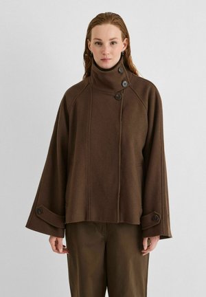 Une femme présente un manteau en laine marron foncé, à col haut boutonné et aux manches larges, sur un fond gris uni.