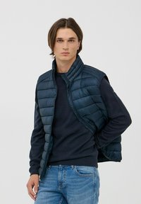 Gilet imbottito blu navy con motivo a costine verticali, colletto alto e zip frontale, indossato sopra un maglione scuro, abbinato a jeans azzurri chiari.