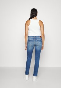 Jeans de mezclilla azul con un corte ajustado, que cuentan con dos bolsillos traseros y un parche de cuero marrón con el logo. Se llevan con una camiseta sin mangas blanca y zapatillas blancas.