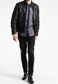 Zwarte leren motorjack met gewatteerde textuur op de schouders, ritsluiting, gedragen over een grijs overhemd met knopen en zwarte jeans.