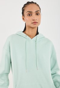 Gaismi piparmētru zaļš džemperis, izgatavots no mīksta auduma. Aprīkots ar regulējamu kapuci, kangaru kabatu un ribotām piedurknēm. Vienkāršs dizains ar brīvu piegriezumu.